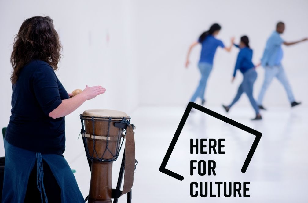 On the left side of the image, a female musician plays a traditional African djembe drum. In the back ground three dancers dressed in blue perform a dance together. The funders logo, here for culture, is on the bottom right of the photo.